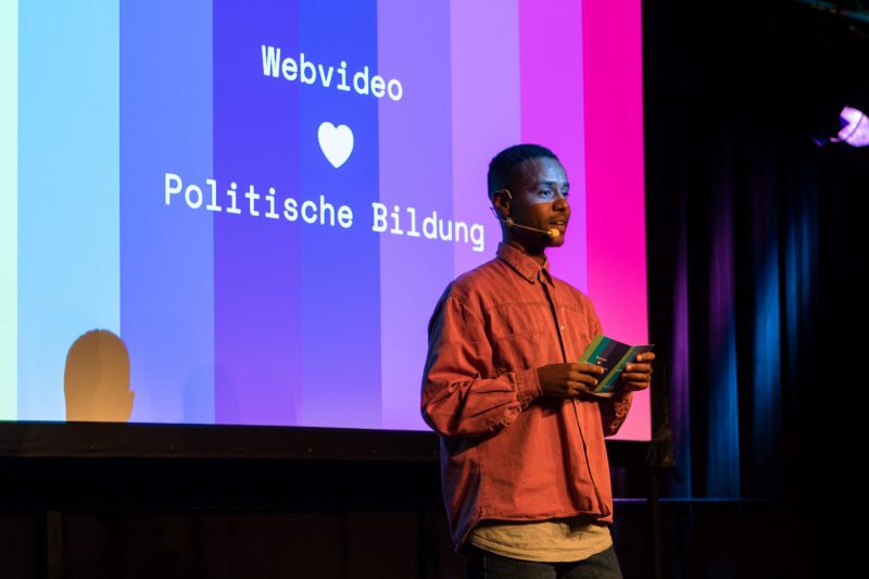 Tarik Tesfu, YouTuber, hält einen Vortrag auf der Fachtagung Bewegtbildung 2018 vor dem Logo der Fachtagung, auf dem steht "Webvideo, gefolgt von einem Herz, gefolgt von den Worten "politische Bildung"