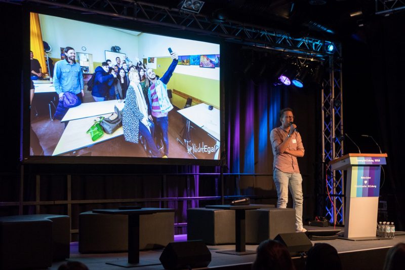 Björn Schreiber von der FSM steht auf der Bühne der Fachtagung Bewegtbildung 2018, hinter ihm ist ein Foto auf der Leinwand, auf der Younis von den Datteltätern ein Selfie mit einer Schüler*innengruppe im Rahmen des Projektes #NichtEgal macht