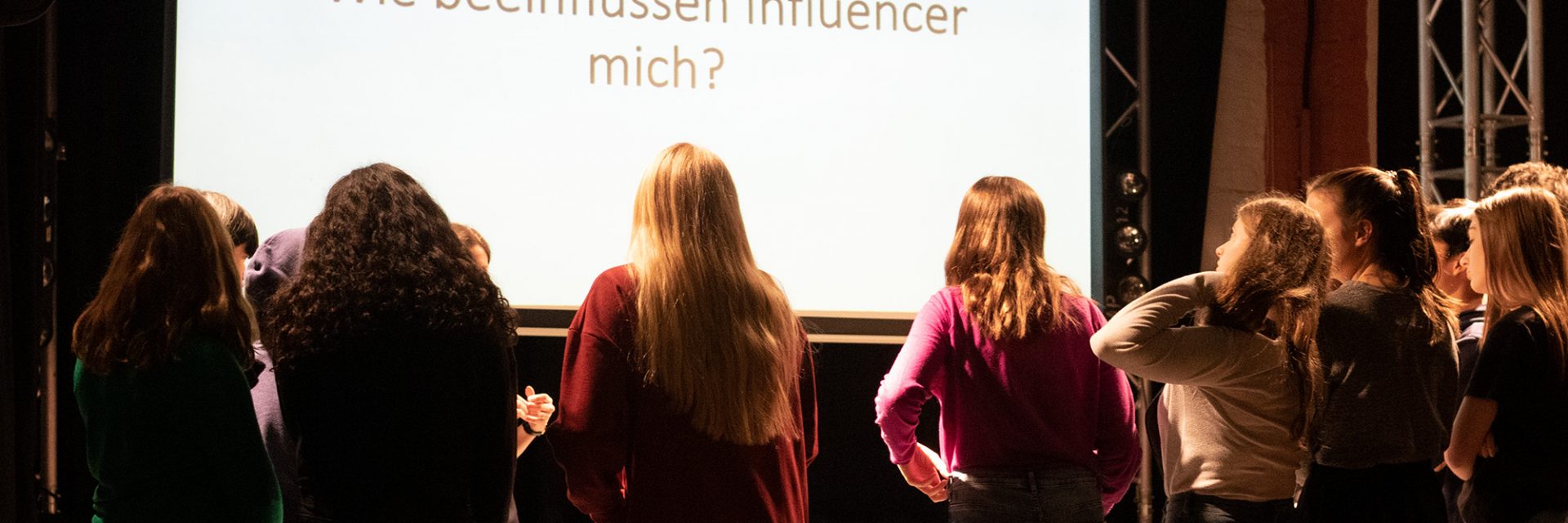 Sechs Mädchen stehen mit dem Rücken zur Kamera und schauen auf eine Leinwand, auf der die Frage "Wie beeinflussen Influencer mich" steht.