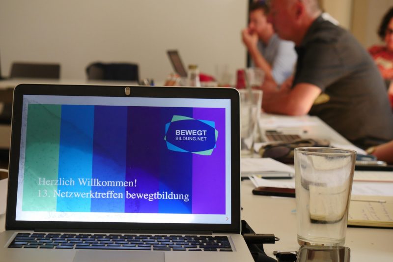Computerbildschirm zeigt das Bewegtbildungslogo und den Text "Herzlich willkommen! 15. Netzwerktreffen Bewegtbildung. Im Hintergrund sieht man zwei Menschen an einem Konferenztisch sitzen.