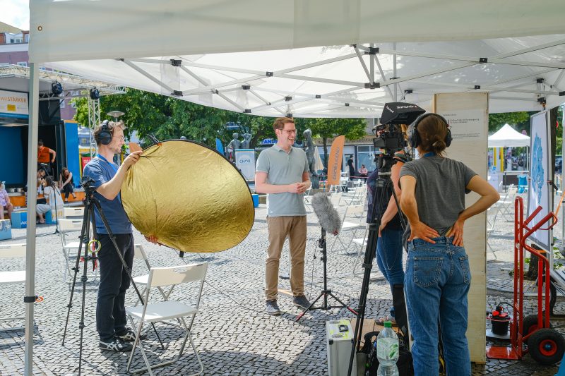 Frau hinter einer Kamera filmt ein Interview zwischen einem Mann und einer Frau, ein weiterer Mann hält eine Sonnenblende.
